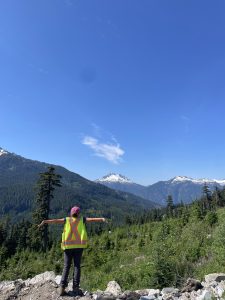 A field crew member standing in front of snow-capped mountains with her back to the camera and her arms outstretched.